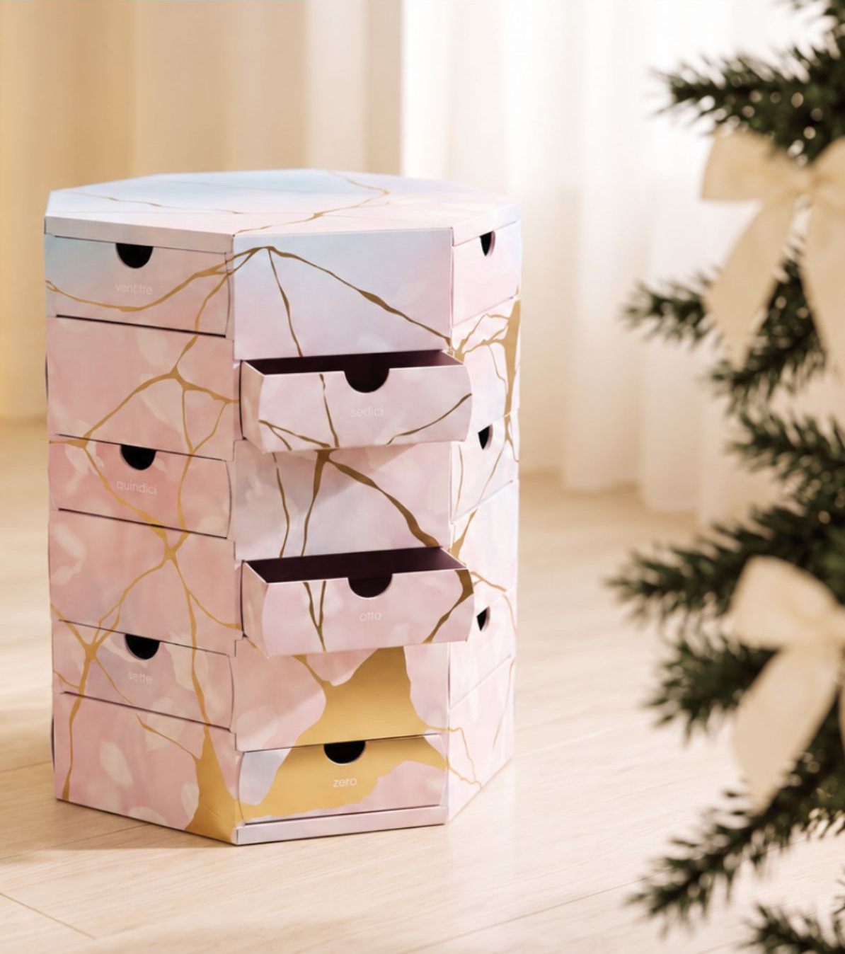 Il calendario ALTROVE intero, esagonale, rosa con venature dorate e i cassetti «sedici» e «otto» aperti, accanto a un albero di Natale con fiocchi color crema